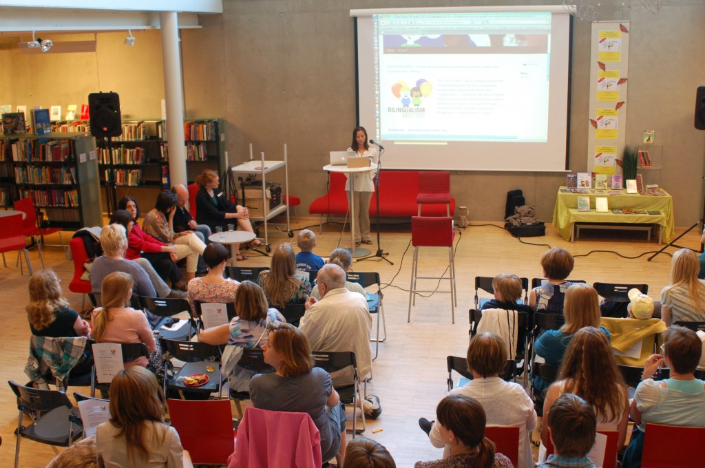 Many people sitting and watching a woman giving a presentation.