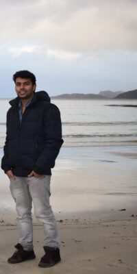 An Indian man wearing a jacket and jeans on the beach