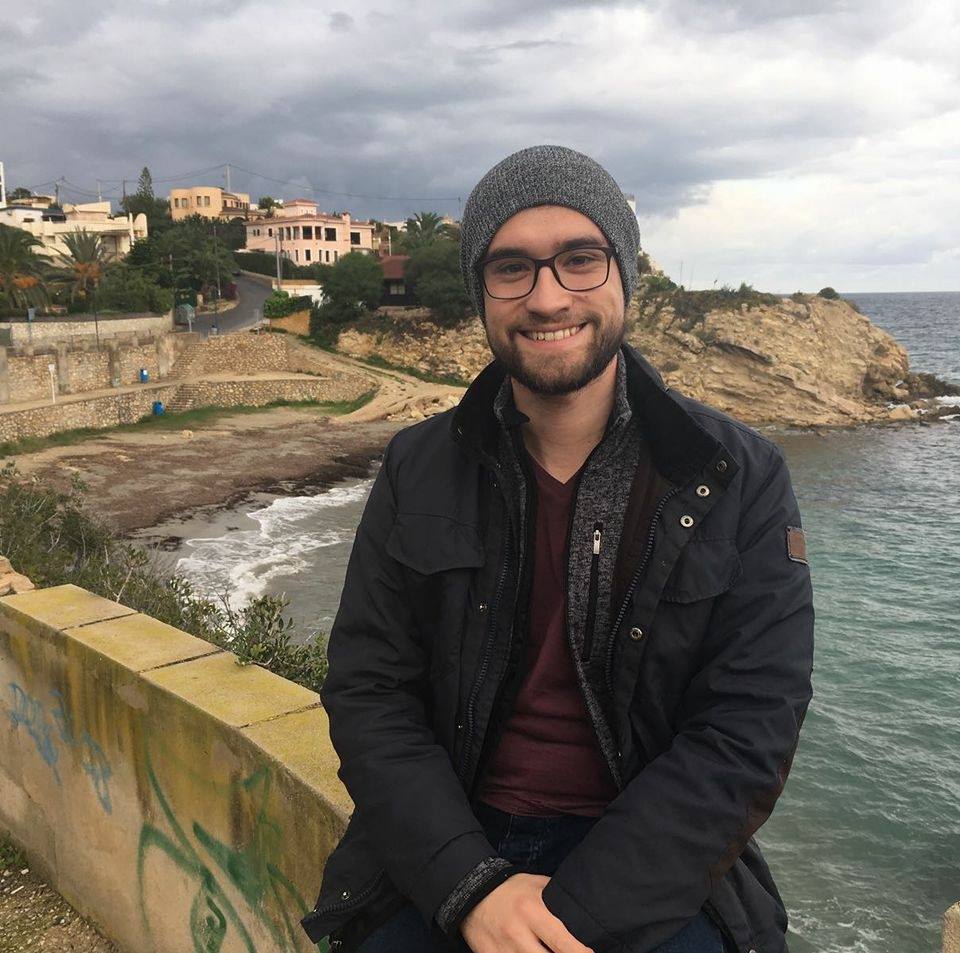 A smiling white man wearing glasses, a short dark beard, and winter cap sitting outside a coastal village