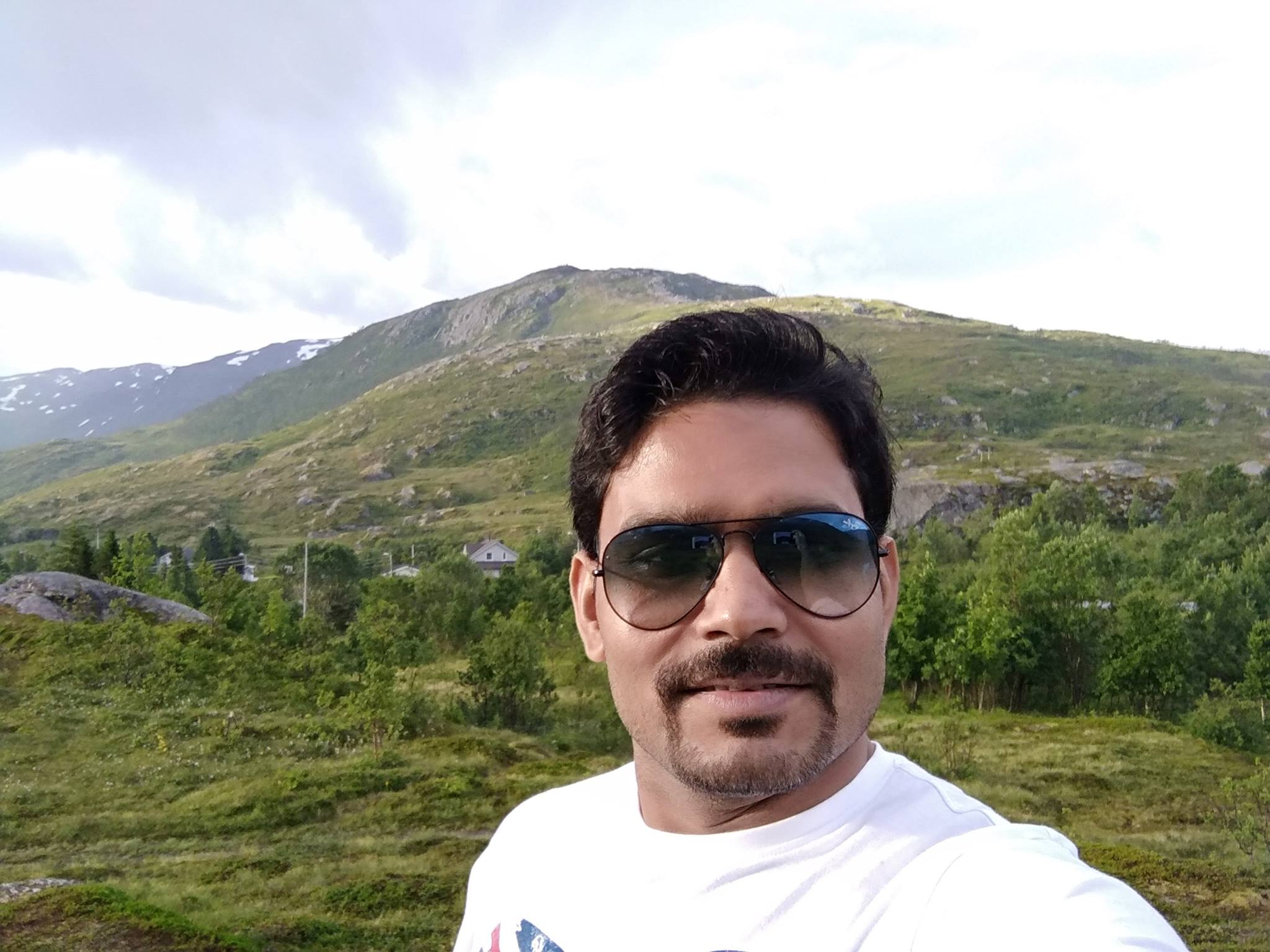 An Indian man with a well-groomed mustache and beard wearing sunglasses in front of a mountain
