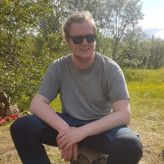 A white man with short blond hair and short beard wearing sunglasses sitting in nature