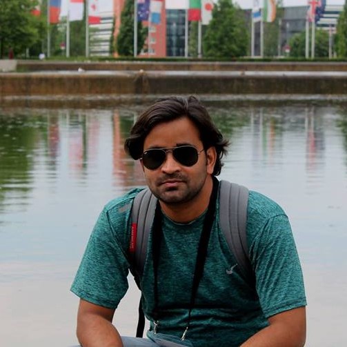 A young Indian man with medium/length hair and sunglasses sitting in front of a river