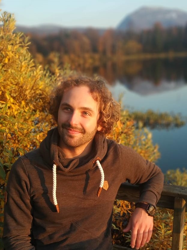 A young smiling white man with medium length curly brown hair sitting in nature 
