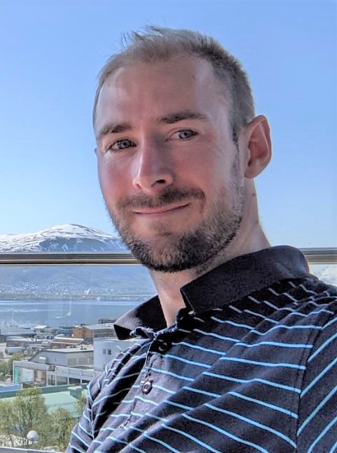 Smiling white man with short hair and a beard in a striped shirt in front of water and a mountain