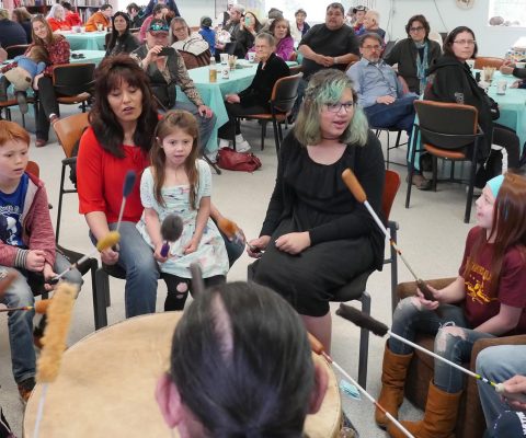Group of children and adults singing and drumming with audience of parents.